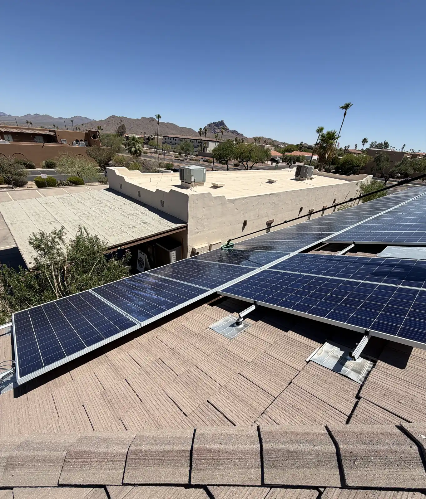 A long array of clean solar panels on a commercial tile roof beside a flat-roof stucco building with rooftop HVAC units, with Sonoran mountains and palm trees behind