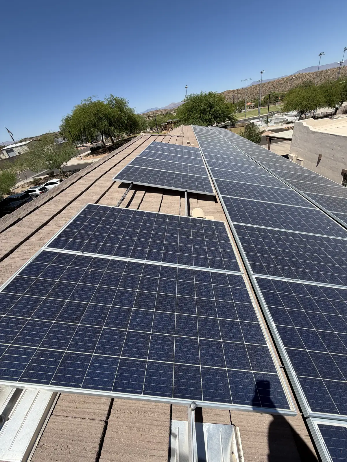 Rows of clean solar panels on the tile roof of a commercial complex, with a sports field, light poles, and desert mountains in the background