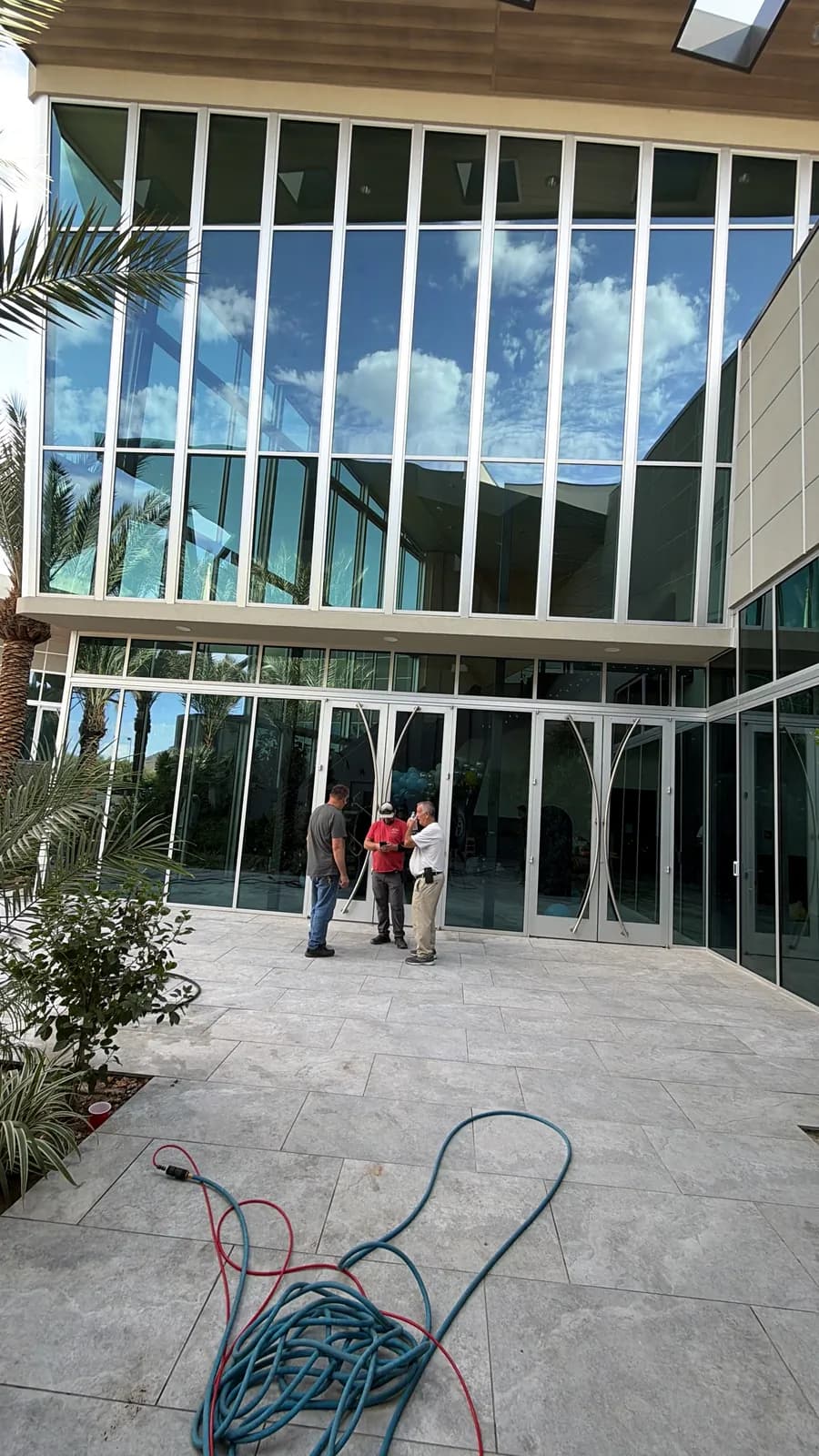 Two-story glass curtain wall and entry doors on a modern commercial building, with three men talking on the stone plaza and a coiled hose in the foreground during a window cleaning job in the Phoenix Valley