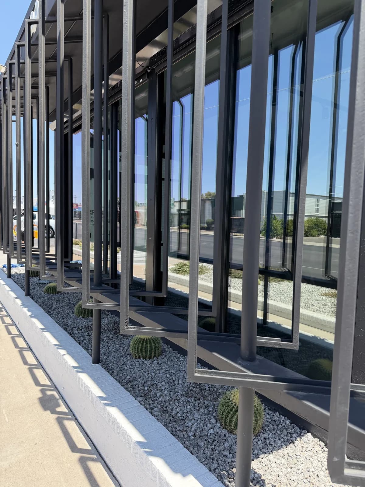 Floor-to-ceiling glass storefront on a modern commercial building, set behind decorative dark steel fins, with a gravel desert planter of barrel cactus in front and a sunny street reflected in the glass