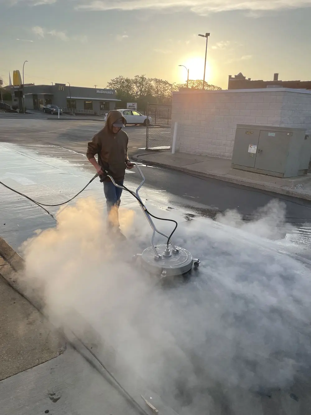 Pressure washing professional using a surface cleaner on a commercial lot at sunset